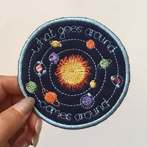 May include: Embroidered patch with a blue background and a light blue border. The patch features a solar system with a yellow sun and colorful planets. The text "What goes around comes around" is embroidered in white on the patch.