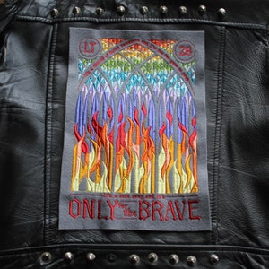 May include: Embroidered patch with a gray background and colorful flames. The text "It's a cold world and it's ONLY for the BRAVE" is embroidered in red.