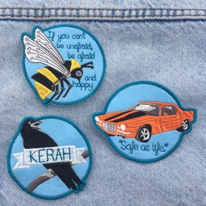 May include: Three embroidered patches on denim. One features a bee with the text "If you can't be unafraid, be afraid and happy." Another shows an orange car with black stripes and the text "Safe as life." The third depicts a crow with the name "KERAH."