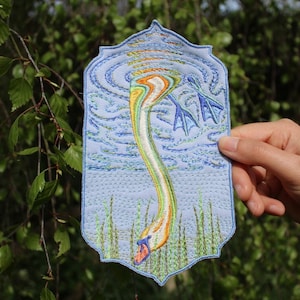 Könnte beinhalten: Gestickter Aufnäher mit einem Schwan mit langem, gebogenem Hals und bunten Federn. Der Schwan ist vor einem hellblauen Hintergrund mit Wasser und Schilf dargestellt. Der Aufnäher wird von einer Hand gehalten, mit einem grünen und blauen Rand.