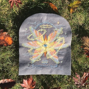 Op de afbeelding: Een grijs vilt patch met een geborduurd ontwerp van een geel en oranje gevleugeld wezen met een blauw oog. Het wezen is omringd door een blauw en wit ontwerp met sterren.