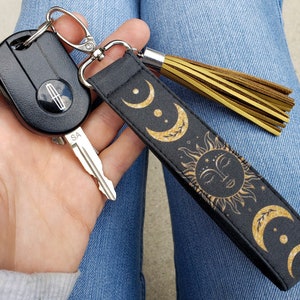 Puede incluir: Un llavero negro con un dise&ntilde;o de sol y luna dorado. El llavero tiene un borla y un anillo de llave plateado.
