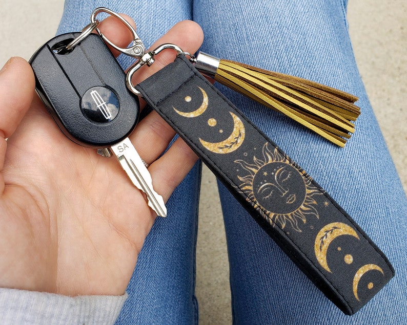 Puede incluir: Un llavero negro con un dise&ntilde;o de sol y luna dorado. El llavero tiene un borla y un anillo de llave plateado.