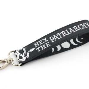 K&ouml;nnte beinhalten: Schwarzer Schl&uuml;sselanh&auml;nger mit einem silbernen Clip und einem wei&szlig;en Schlangenmuster. Der Schl&uuml;sselanh&auml;nger tr&auml;gt den Text "Hex the Patriarchy" und ein Design mit Halbmond und Stern.
