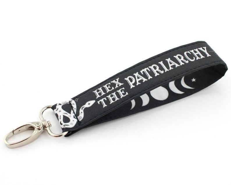K&ouml;nnte beinhalten: Schwarzer Schl&uuml;sselanh&auml;nger mit einem silbernen Clip und einem wei&szlig;en Schlangenmuster. Der Schl&uuml;sselanh&auml;nger tr&auml;gt den Text "Hex the Patriarchy" und ein Design mit Halbmond und Stern.