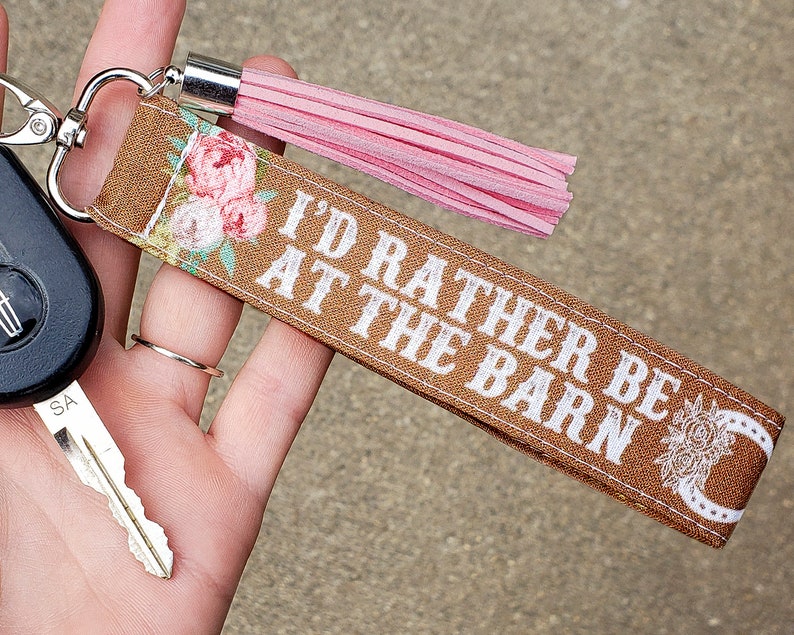 May include: Brown fabric key chain with a pink tassel. The key chain has white text that reads "I'd rather be at the barn" and a white horseshoe design.