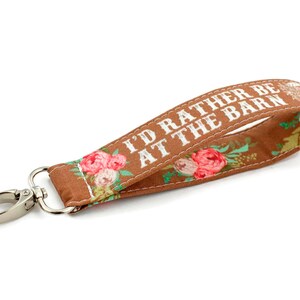 May include: Brown fabric key chain with white text that reads "I'd rather be at the barn" and floral print. The key chain has a silver metal clasp.