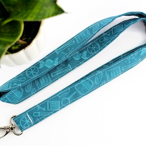 Könnte beinhalten: Türkisfarbenes Stoff-Lanyard mit einem wissenschaftlichen Muster mit Laborausrüstung, DNA-Strängen und Atomsymbolen. Das Lanyard hat einen silberfarbenen Metallverschluss. Der Stoff ist mit weißem Faden genäht.