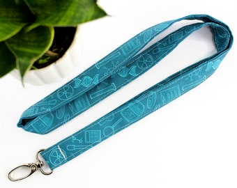 Op de afbeelding: Teal stoffen lanyard met een wetenschappelijk thema patroon met laboratoriumapparatuur, DNA-strengen en atoom symbolen. De lanyard heeft een zilverkleurige metalen sluiting. De stof is gestikt met witte draad.