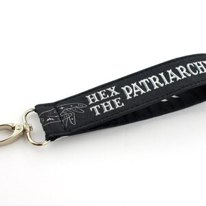 K&ouml;nnte beinhalten: Schwarzer Schl&uuml;sselanh&auml;nger mit einem silbernen Clip und dem Text "Hex the Patriarchy" in wei&szlig;en Buchstaben.