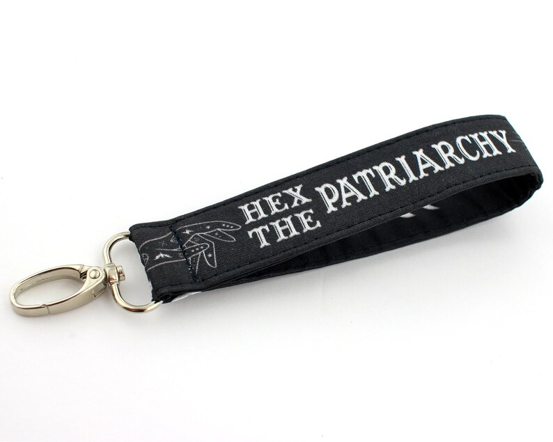 K&ouml;nnte beinhalten: Schwarzer Schl&uuml;sselanh&auml;nger mit einem silbernen Clip und dem Text "Hex the Patriarchy" in wei&szlig;en Buchstaben.