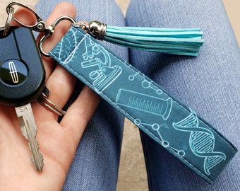 May include: Teal blue fabric keychain with a light blue tassel. The keychain features a microscope, a beaker, and a DNA strand design.