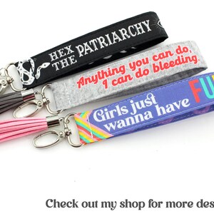 K&ouml;nnte beinhalten: Drei bunte Schl&uuml;sselanh&auml;nger mit Quasten. Der erste Schl&uuml;sselanh&auml;nger ist schwarz mit wei&szlig;em Text, der "Hex the Patriarchy" lautet. Der zweite Schl&uuml;sselanh&auml;nger ist grau mit wei&szlig;em Text, der "Anything you can do, I can do bleeding." lautet. Der dritte Schl&uuml;sselanh&auml;nger ist lila mit Regenbogenstreifen und wei&szlig;em Text, der "Girls just wanna have FUN" lautet.