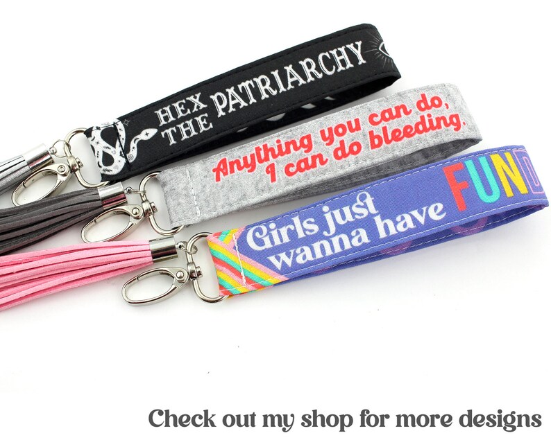 K&ouml;nnte beinhalten: Drei bunte Schl&uuml;sselanh&auml;nger mit Quasten. Der erste Schl&uuml;sselanh&auml;nger ist schwarz mit wei&szlig;em Text, der "Hex the Patriarchy" lautet. Der zweite Schl&uuml;sselanh&auml;nger ist grau mit wei&szlig;em Text, der "Anything you can do, I can do bleeding." lautet. Der dritte Schl&uuml;sselanh&auml;nger ist lila mit Regenbogenstreifen und wei&szlig;em Text, der "Girls just wanna have FUN" lautet.