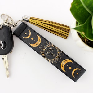 Puede incluir: Llavero negro con un dise&ntilde;o de sol y luna dorado y un borla dorada. El llavero est&aacute; unido a una llave de coche.