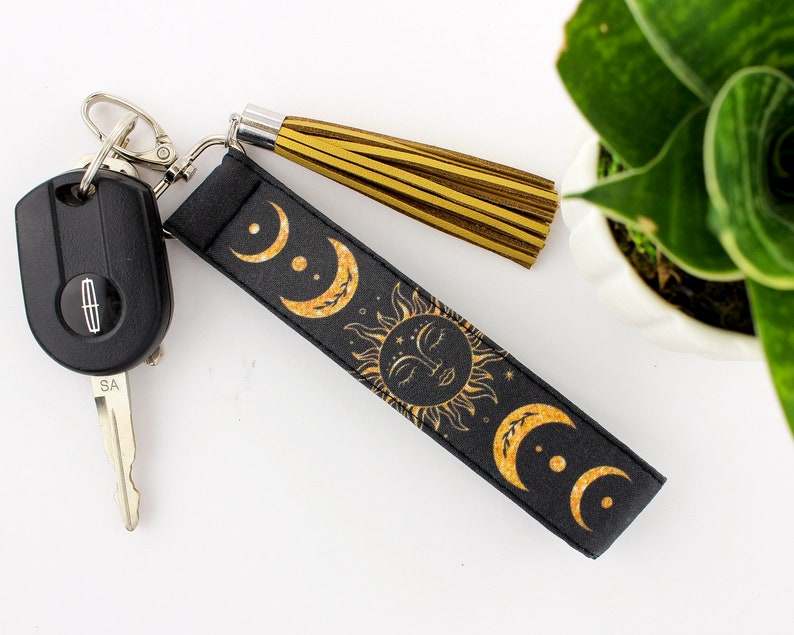 Puede incluir: Llavero negro con un dise&ntilde;o de sol y luna dorado y un borla dorada. El llavero est&aacute; unido a una llave de coche.