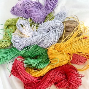 Puede incluir: Una colecci&oacute;n de carretes de hilo de bordar de colores en tonos de morado, verde, amarillo, rojo y marr&oacute;n. Los carretes est&aacute;n dispuestos en una pila suelta sobre una superficie blanca.