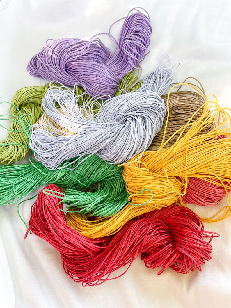 Puede incluir: Una colecci&oacute;n de carretes de hilo de bordar de colores en tonos de morado, verde, amarillo, rojo y marr&oacute;n. Los carretes est&aacute;n dispuestos en una pila suelta sobre una superficie blanca.