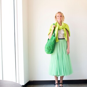 Puede incluir: Una mujer con un su&eacute;ter de punto verde, una blusa blanca con flores, una falda de tul verde y zapatos negros. Lleva un bolso de mano de crochet verde.