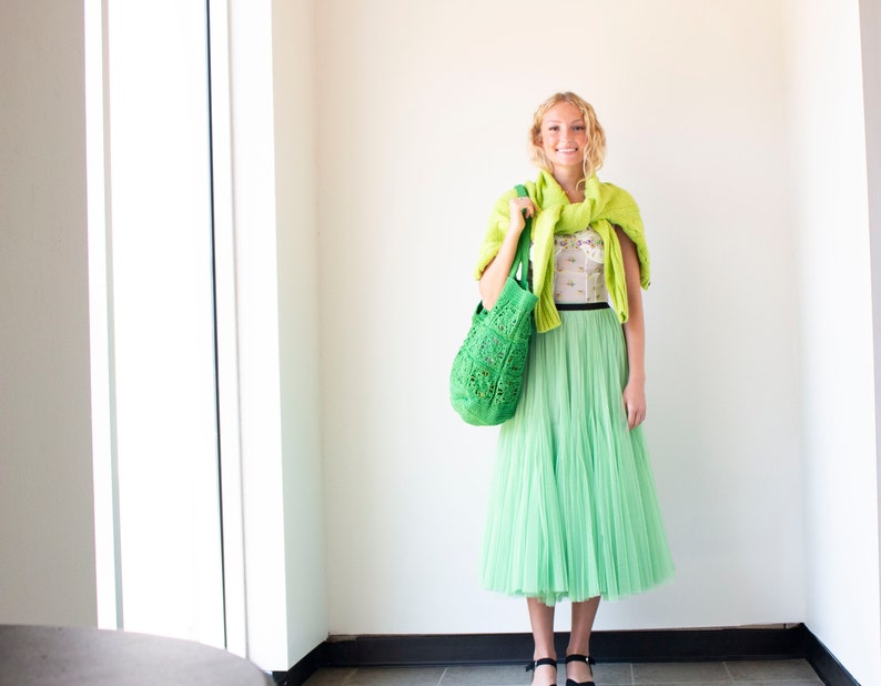 Puede incluir: Una mujer con un su&eacute;ter de punto verde, una blusa blanca con flores, una falda de tul verde y zapatos negros. Lleva un bolso de mano de crochet verde.