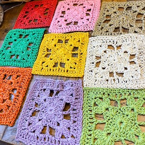 Puede incluir: Nueve cuadrados de ganchillo en varios colores, incluyendo rojo, rosa, amarillo, verde, naranja y morado. Los cuadrados est&aacute;n dispuestos en una fila sobre una superficie de madera.