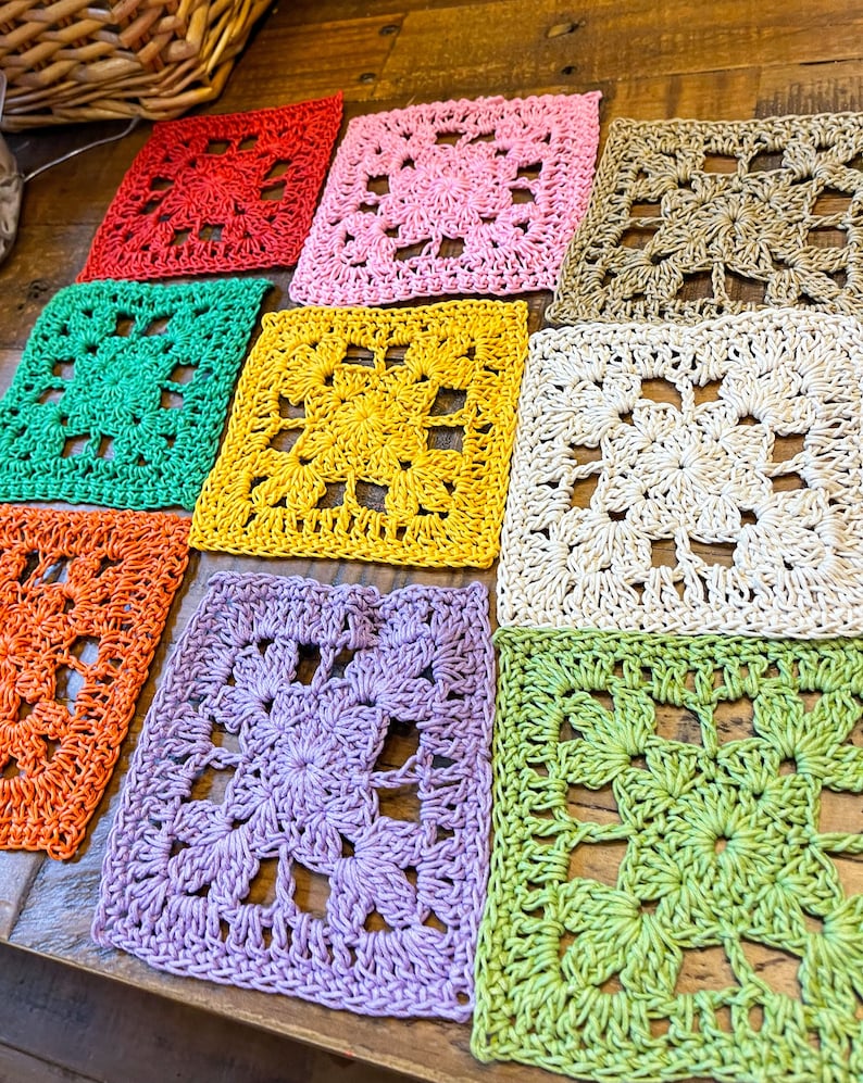 Puede incluir: Nueve cuadrados de ganchillo en varios colores, incluyendo rojo, rosa, amarillo, verde, naranja y morado. Los cuadrados est&aacute;n dispuestos en una fila sobre una superficie de madera.