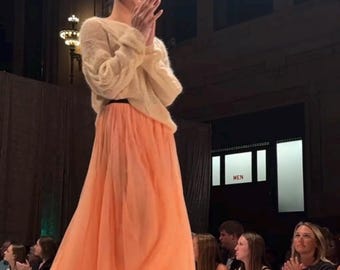 May include: A model on a runway wearing a peach-colored tulle skirt and a cream-colored sweater. The skirt is long and flowing, and the sweater has long sleeves. The model is wearing beige heels and a black belt. The background shows an audience.