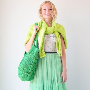 Puede incluir: Una mujer con un su&eacute;ter de punto verde sobre una blusa blanca con estampado floral, una falda de tul verde y una bolsa de mano de crochet verde.
