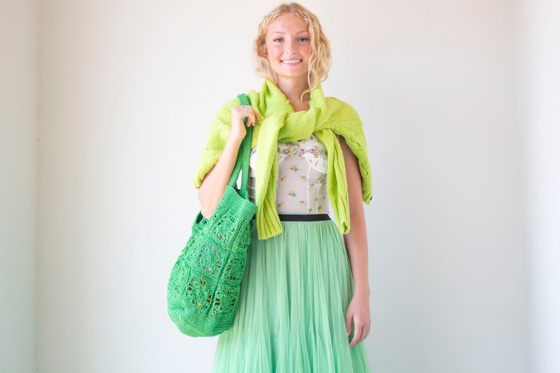 Puede incluir: Una mujer con un su&eacute;ter de punto verde sobre una blusa blanca con estampado floral, una falda de tul verde y una bolsa de mano de crochet verde.