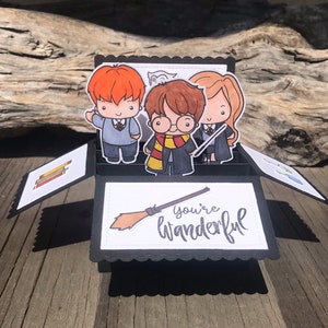 Op de afbeelding: Een pop-upkaart met cartoon illustraties van drie personages met rood, bruin en blond haar, gekleed in schooluniformen en met toverstokken. De kaart heeft de woorden "You're Wonderful" en een bezem illustratie.