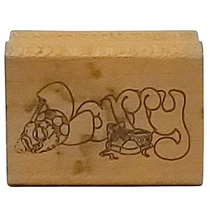May include: A light brown wooden stamp with the word "Dappy" carved into the surface. The design includes a turtle and a frog. The stamp has a rectangular shape with a raised top edge.