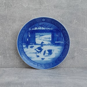 May include: Blue and white Christmas plate featuring a winter scene with a farmhouse, a path, and two geese. The plate has a blue and white border with a star in the centre and the year 1969 printed on the bottom.