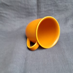 May include: A bright orange ceramic mug with a rounded shape and a handle. The mug is empty and sitting on a gray fabric surface.