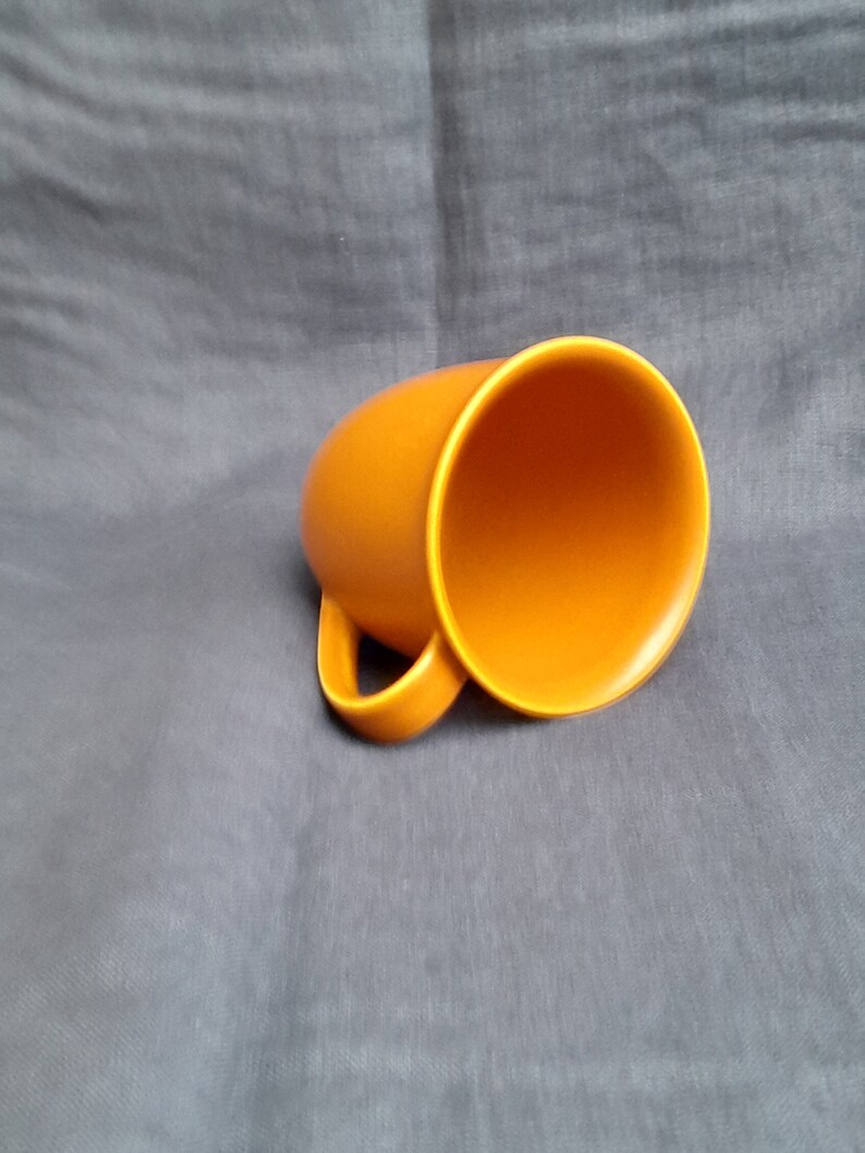 May include: A bright orange ceramic mug with a rounded shape and a handle. The mug is empty and sitting on a gray fabric surface.