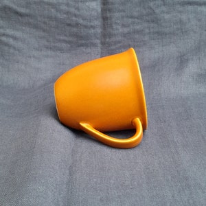 May include: A bright orange ceramic mug with a rounded shape and a handle. The mug is sitting on a gray fabric surface.
