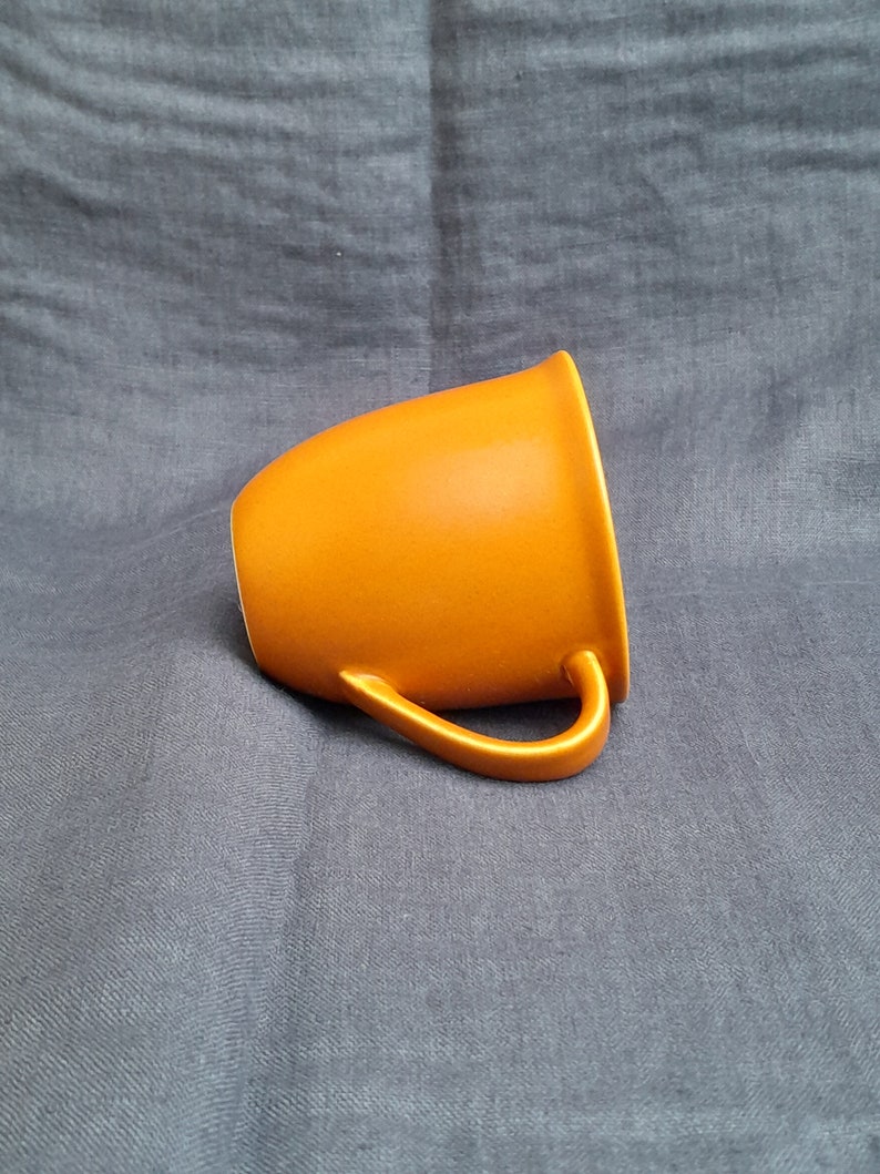 May include: A bright orange ceramic mug with a rounded shape and a handle. The mug is sitting on a gray fabric surface.