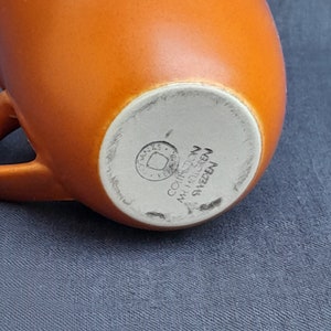 May include: Bottom of an orange ceramic mug with a white mark that reads "COHRE  MADE IN SWEDEN".