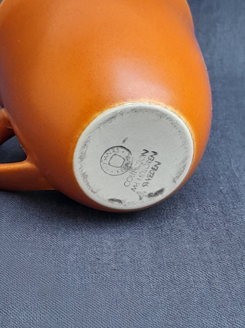 May include: Bottom of an orange ceramic mug with a white mark that reads "COHRE  MADE IN SWEDEN".
