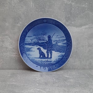 May include: Blue and white ceramic plate depicting a winter scene with a man and his dog. The plate is decorated with a pine tree border and the year 1977.