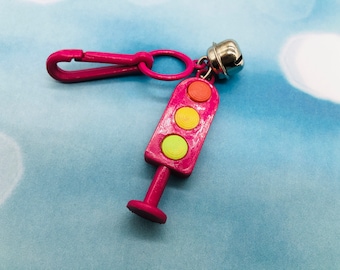 May include: A pink plastic traffic light keyring with a yellow, orange, and red light. The keyring has a silver bell and a pink plastic clip.