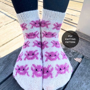 May include: A pair of white knitted socks with a pink crab pattern. The socks are shown on a person's legs. The text "PDF Knitting Pattern" is visible in the image.