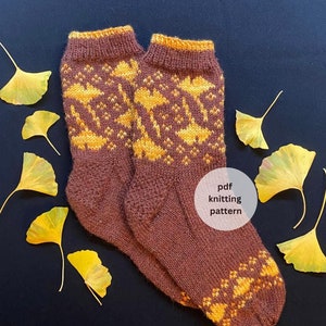 May include: A pair of brown knitted socks with a yellow and orange ginkgo leaf pattern. The socks are on a dark blue background with yellow ginkgo leaves scattered around them. The text "pdf knitting pattern" is written in white on a white circle.