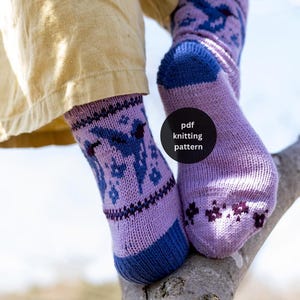 May include: A pair of light purple knit socks featuring a dark blue bird and star pattern, with dark blue cuffs and toes. The socks display a small dark floral motif near the toe area and include the text pdf knitting pattern on the side.
