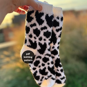 May include: A pair of knitted socks featuring a black and white cow print pattern with a white ribbed cuff and white toe section. A circular black label on the side of the sock displays the white text pdf knitting pattern.