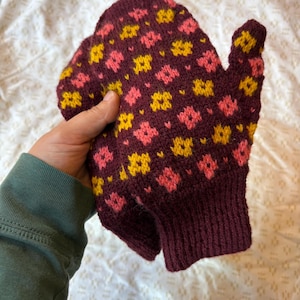 May include: A pair of knitted mittens in a deep burgundy color, adorned with a repeating floral pattern in yellow and pink. The mittens have a ribbed cuff and are held up.