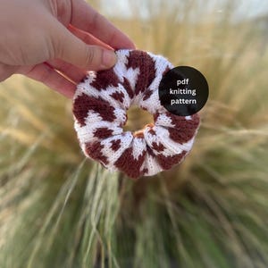 Op de afbeelding: Een gebreide scrunchie met een tweekleurig bruin en wit koeienvlekkenpatroon. Een zwarte cirkelvormige grafische overlay aan de rechterkant bevat de witte tekst pdf knitting pattern. Het accessoire heeft een geplooide, elastische textuur.