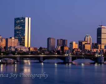 May include: A panoramic view of the Boston skyline at dusk, with the Charles River in the foreground and several bridges connecting the city to the other side. The skyscrapers are illuminated by the setting sun, casting a warm glow over the cityscape.