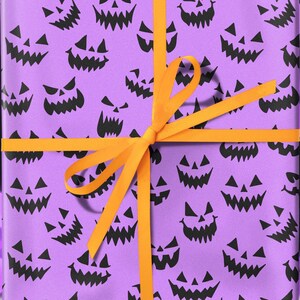 May include: A wrapped gift with a purple background featuring black jack-o'-lantern faces. The gift is tied with an orange ribbon. The background has a bokeh effect with purple lights. The gift is on a light-colored surface.