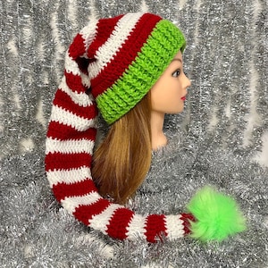 May include: A handmade elf hat with a long, striped tail. The hat features alternating red and white stripes on the tail, a green ribbed knit cap, and a fluffy green pom-pom at the end. The hat is designed for festive occasions.