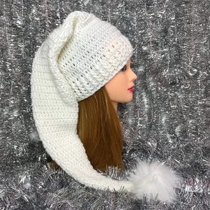 May include: A white, crocheted hat with a long, pointed end and a fluffy white pom-pom. The hat is displayed on a mannequin head with long brown hair, against a silver tinsel background. This whimsical hat is perfect for costume parties or festive occasions.